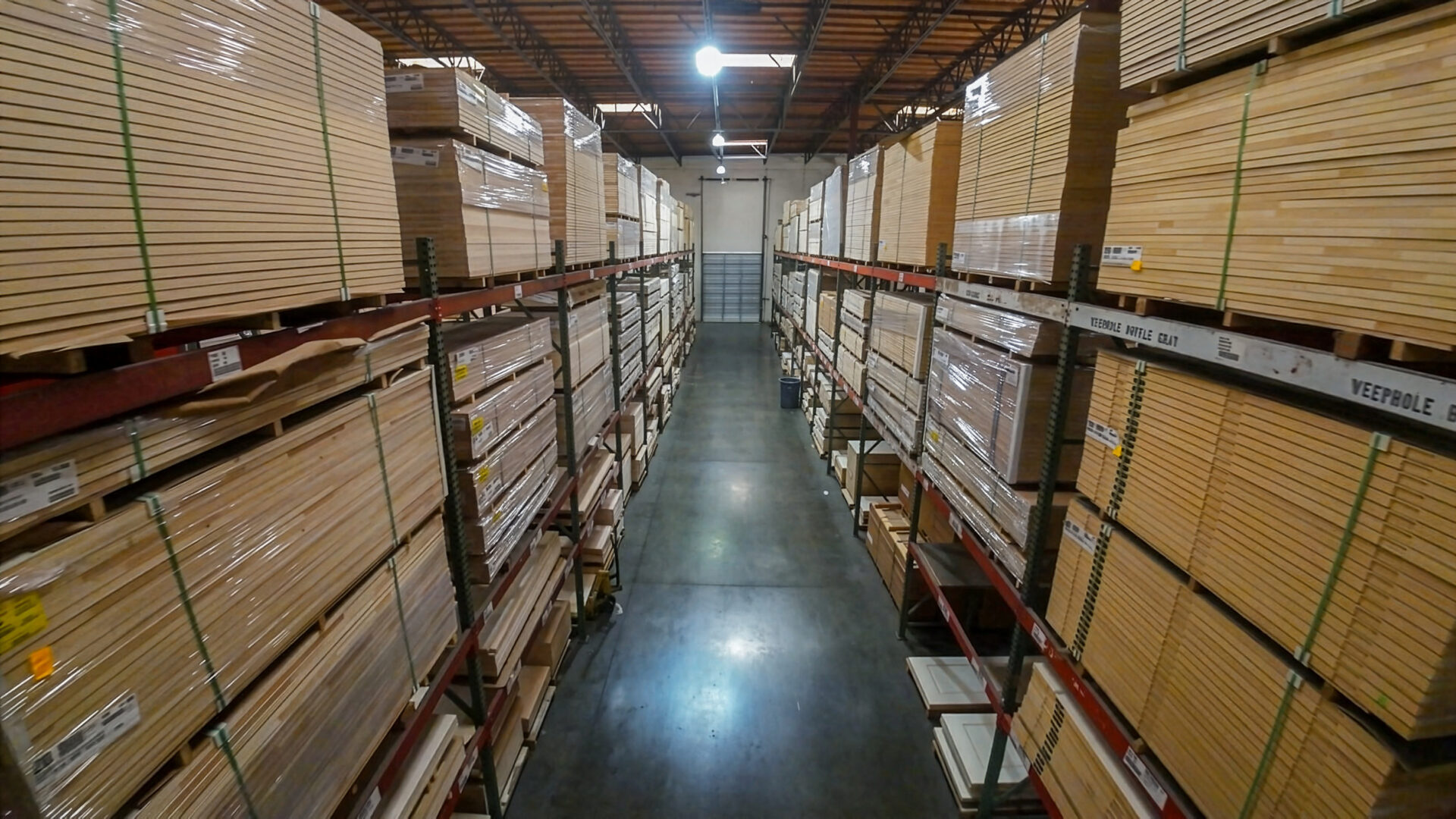 A long warehouse aisle with tall racks of stacked plywood and lumber on both sides, pallets wrapped in plastic.